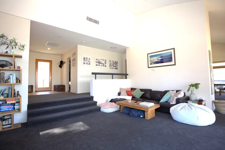 Bright, open living area with a dark sectional sofa and colorful cushions, two white bean bags, a wooden coffee table, and a bookshelf on the left; steps and a hallway lead to another room, framed art on the wall, and potted plants nearby.