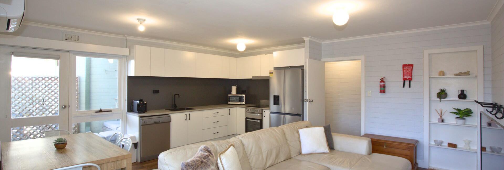 Open-plan living room and kitchen with white cabinets, stainless-steel appliances, and a beige sectional sofa against a light gray wall.