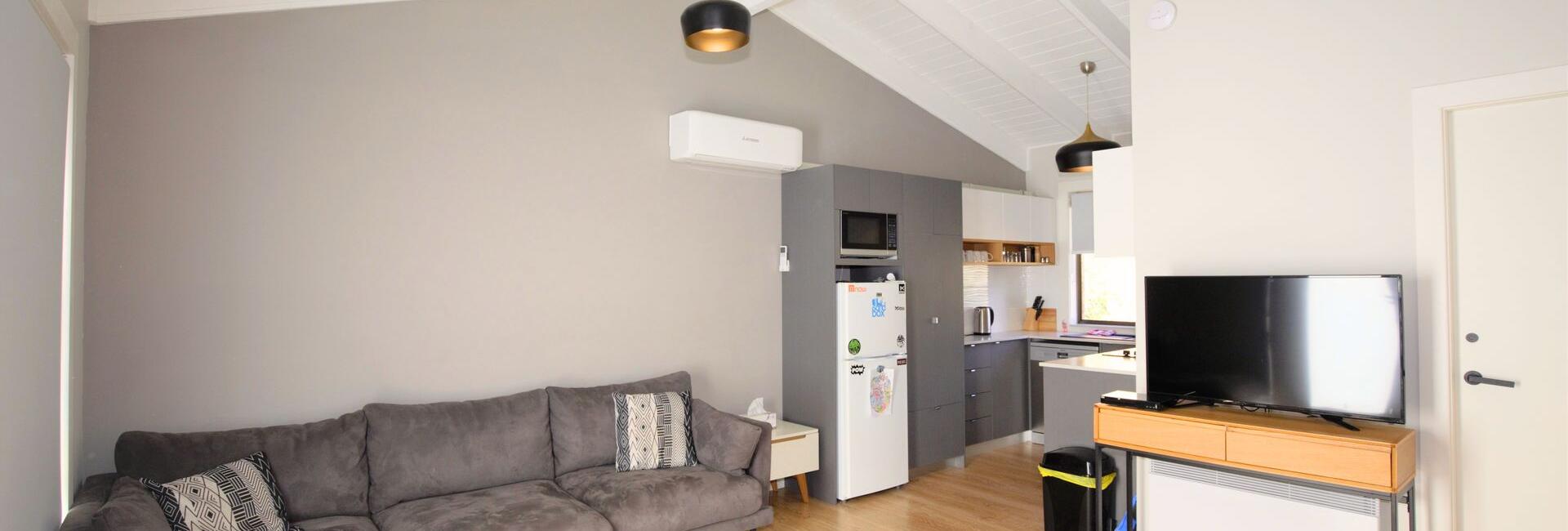 Open-plan living area with a grey sectional sofa, white vaulted ceiling with exposed beams, and a kitchen area with cabinets and fridge visible nearby.