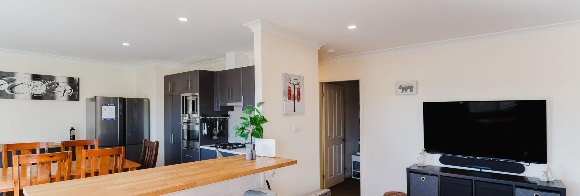 Open-plan kitchen and living area with a wooden bar counter and three bar stools, and a wall-mounted TV on the right.
