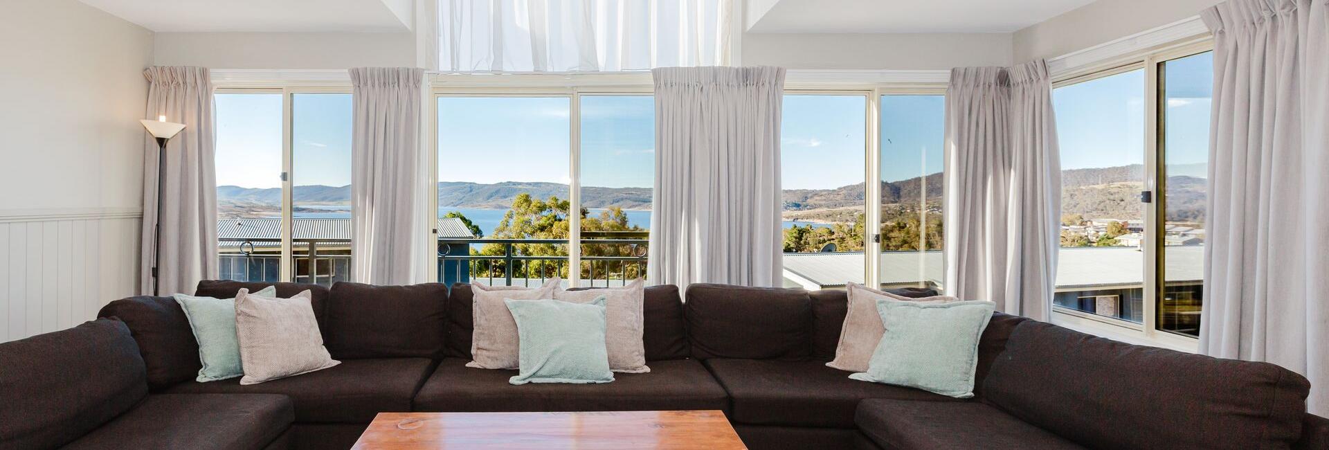 Living room with a large brown sectional, light pillows, and a wooden coffee table, facing big windows with a lake view.