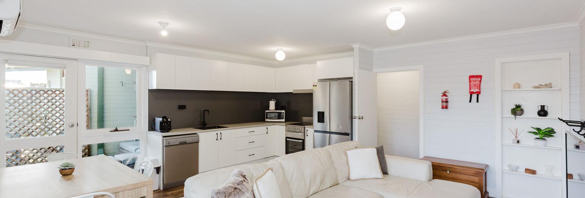 Open-plan living room and kitchen with white cabinets, stainless appliances, beige sectional sofa, and light wood floors.