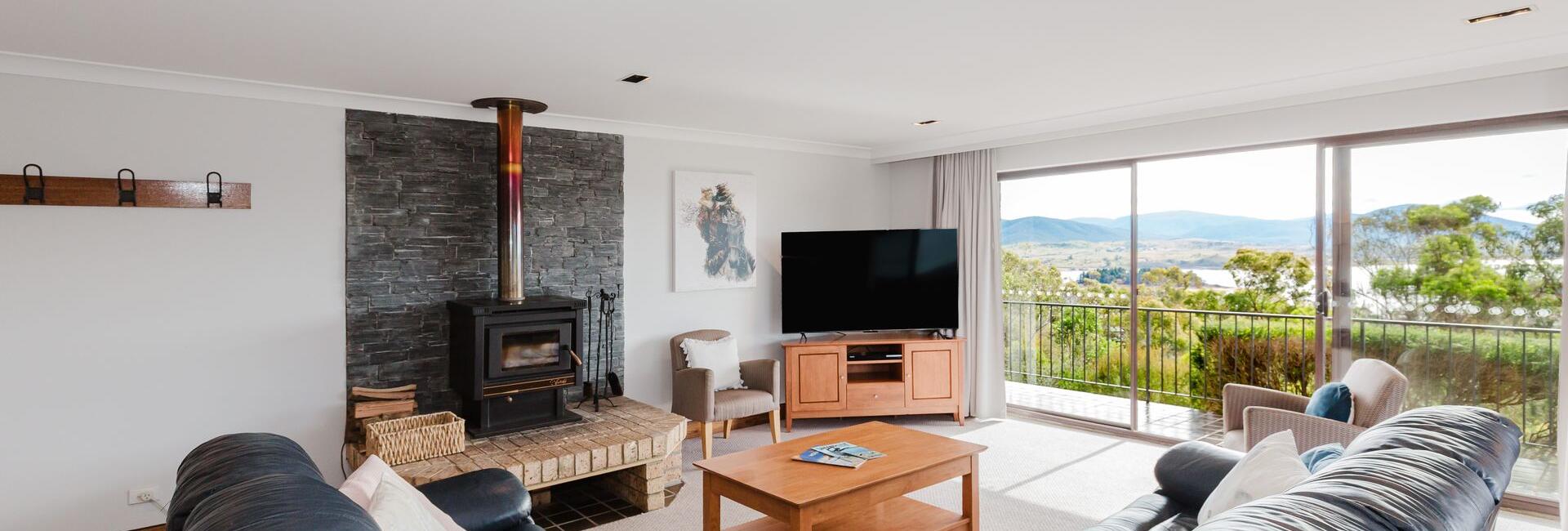 Spacious living room with a stone fireplace and wood-burning stove, a dark leather sectional, and a wooden coffee table facing a flat-screen TV by large glass doors to a balcony.
