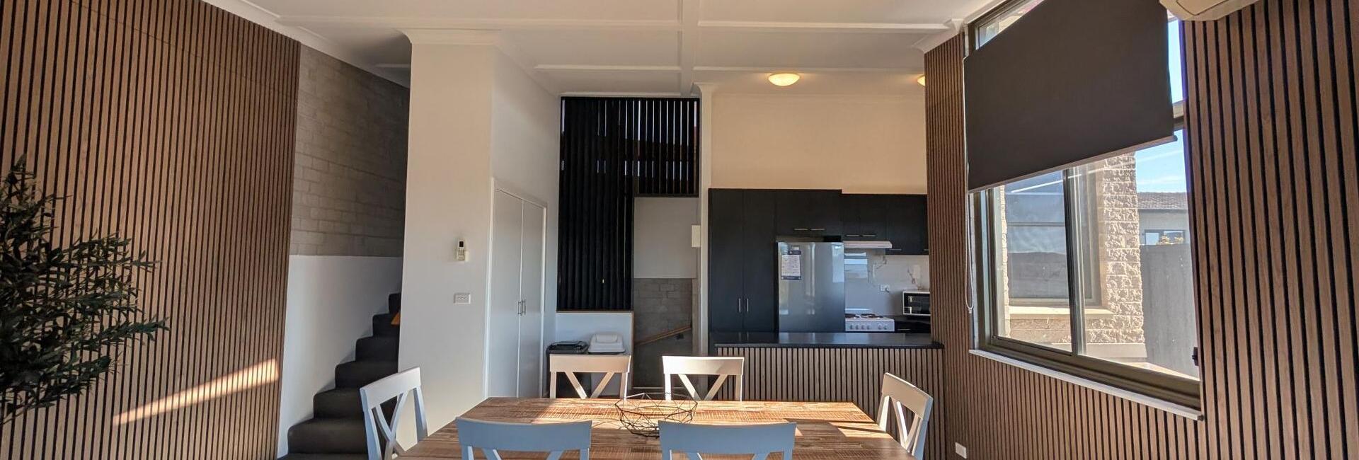 Open-plan dining area with a wooden slat wall, a rectangular table and white chairs, and a modern kitchen in the background.